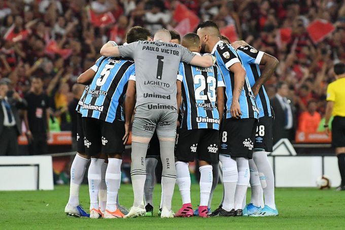 a grupo de jogadores do grêmio abraçados, em circulo, de costas para a câmera