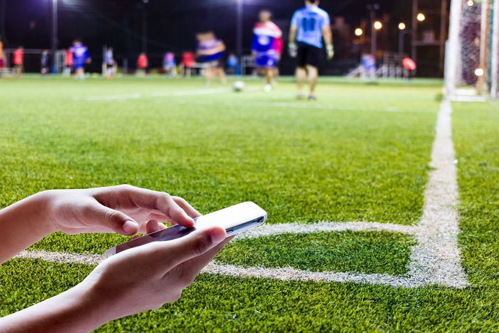 Uma mão, sobre um campo de futebol, usa o celular