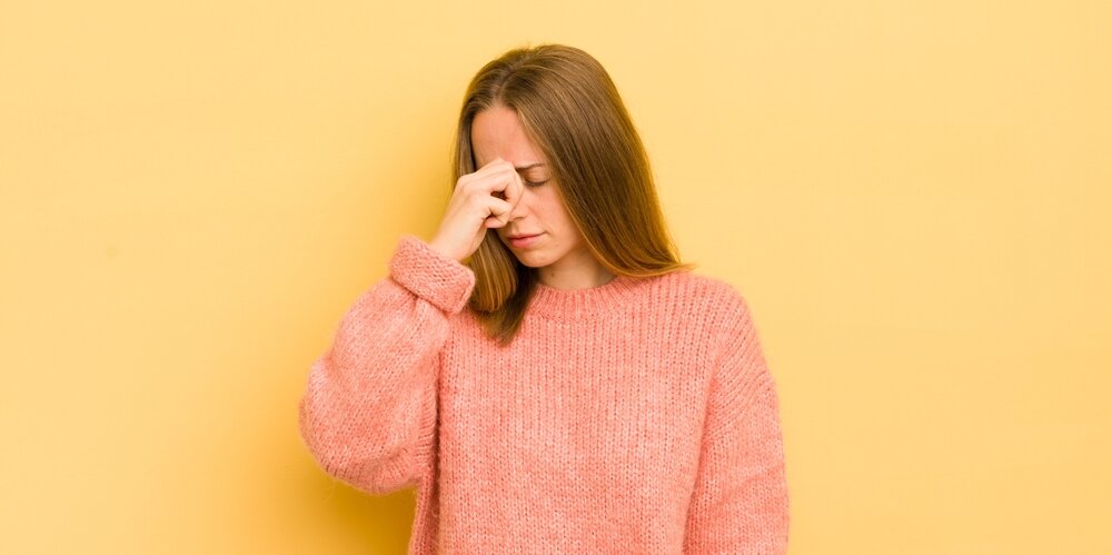 A young woman in a pink jumper pinching her forehead, standing in front of a yellow wall.