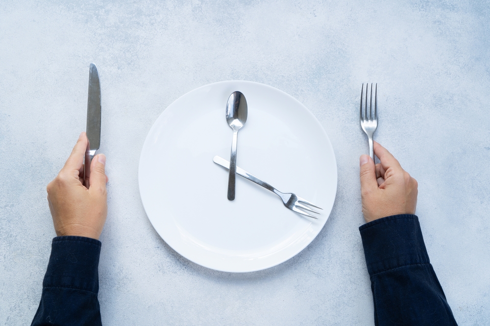 A white plate with spoon and fork in shape of a clock, and two hands with a knife and fork alongside it