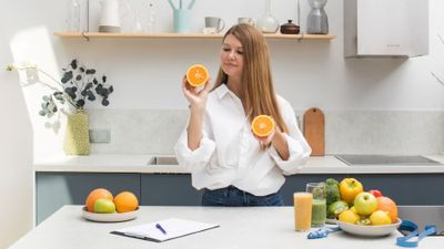 Woman in kitchen  holding two sides of orange cut in half.  Various fruit and vegetables on counter.