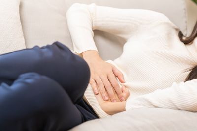 A woman laying on a couch with her knees pulled up towards her chest and hands pressed against her stomach.