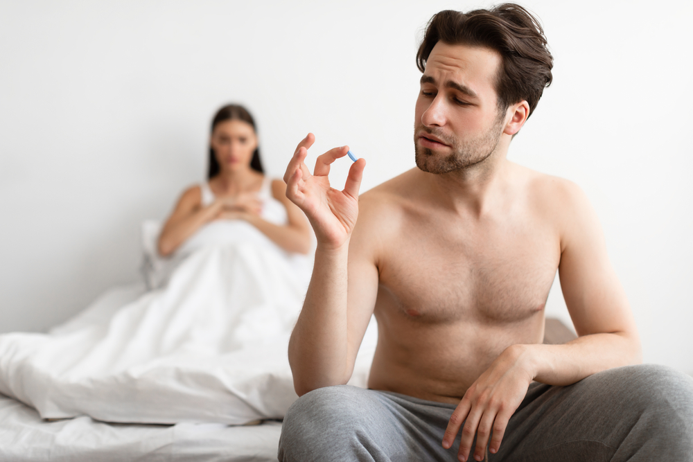 Man sitting on the bed holding a pill with woman under the bed covers in the background