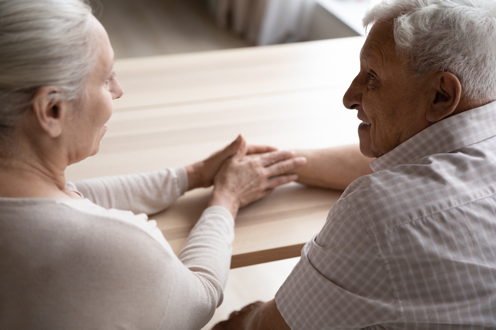 Elderly couple sitting at a table together while holding hands and smiling gently