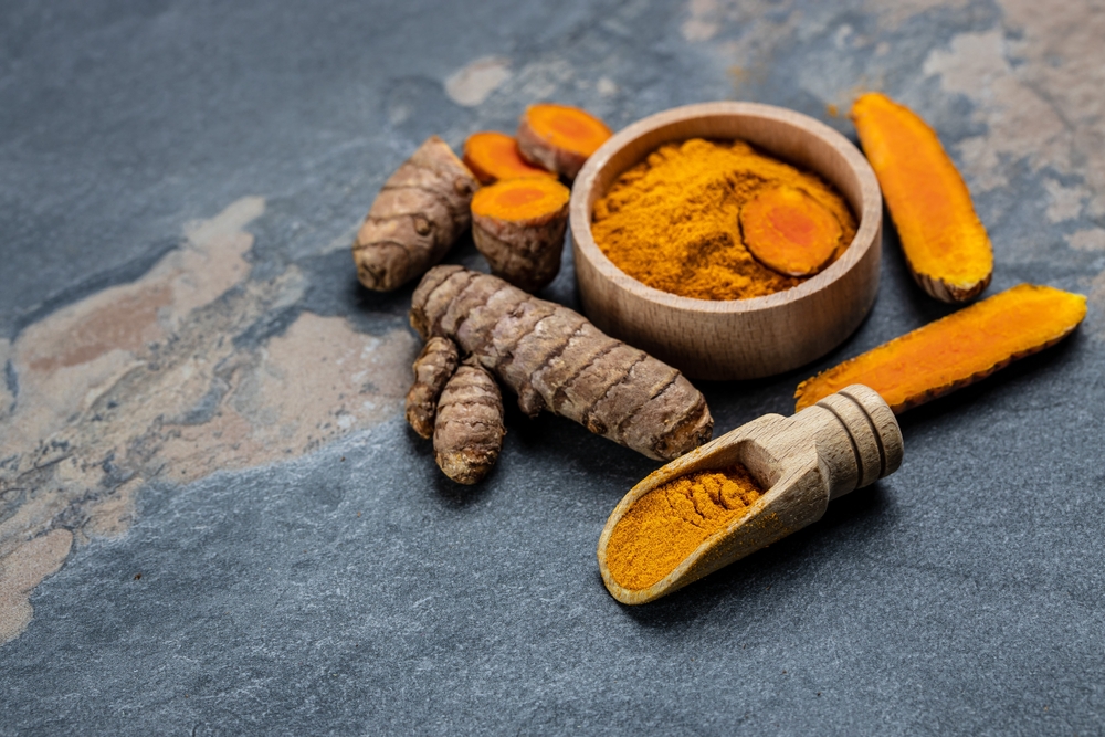 Turmeric powder in a bowl surrounded by fresh turmeric on a dark surface