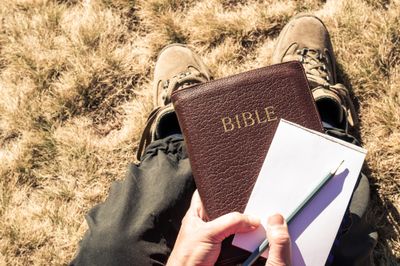 Man outdoors with hiking boots and trousers holding a Bible with pencil and notepad