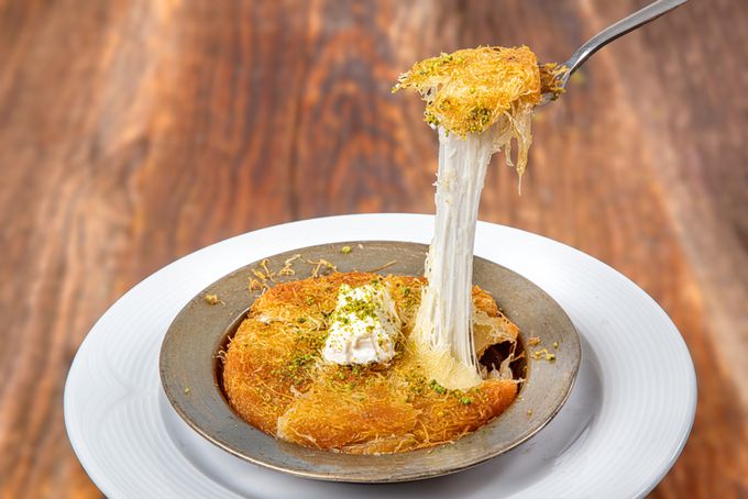 A bowl of Knafeh topped with pistachios and a piece being pulled up with spoon  