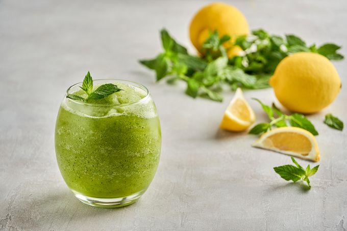 A tumbler with green beverage inside topped with mint, surrounded by mint and lemons