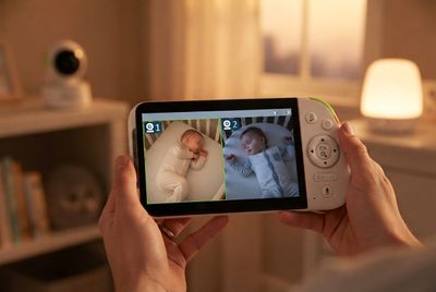 A parent holding a Babysense PRO parent unit in a softly lit nursery, with two baby camera views shown side by side on the screen
