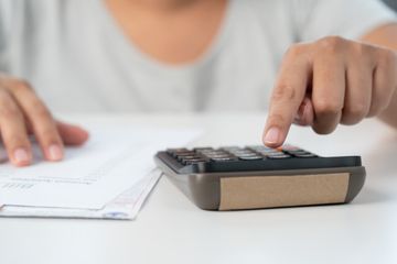 A person seated at their desk, busy punching numbers from a page in front of them into a calculator.