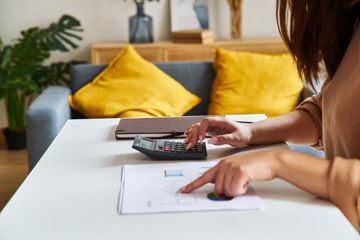 A woman seated at her living room table, performing calculations while reviewing something on paper.
