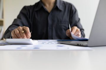 A man seated at a desk in his office, performing a calculation, with a laptop and various papers visible on his desk.