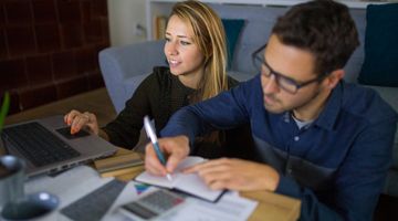 Man writing in notebook while blonde woman looks at laptop screen