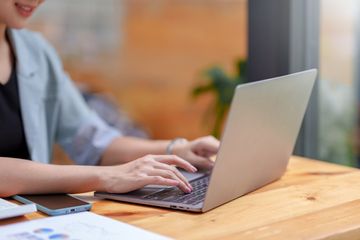 A woman seated at a desk, working on her laptop, with her phone, a notebook, and stack of papers placed next to her.