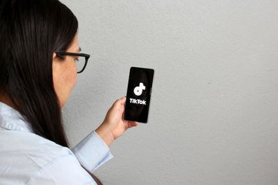 A woman looking at her cellphone in her hand, displaying the TikTok logo in white against a black background.