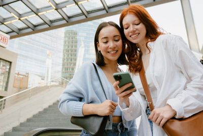 Two young woman standing together, smiling as the one shows the other something on her cellphone.