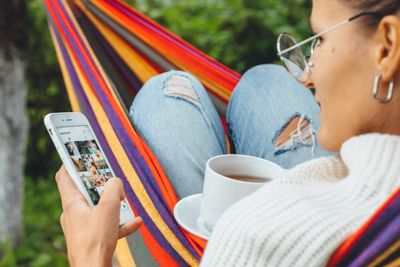 A woman, seated with a cup of tea in a hammock outside, scrolling through Instagram.