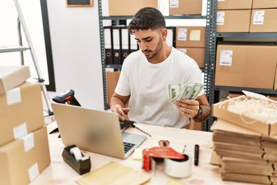 Person using laptop and holding cash surrounded by boxes
