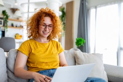 A young woman with red hair, wearing a yellow shirt, seated in her living room, working on her laptop.
