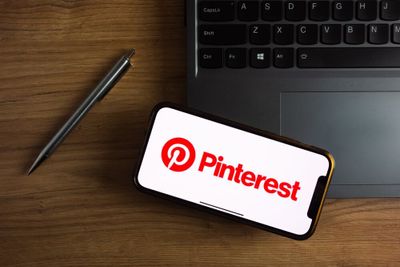 An iPhone screen displaying a white background with Pinterest's logo in red, along with a laptop and pen on a desk.