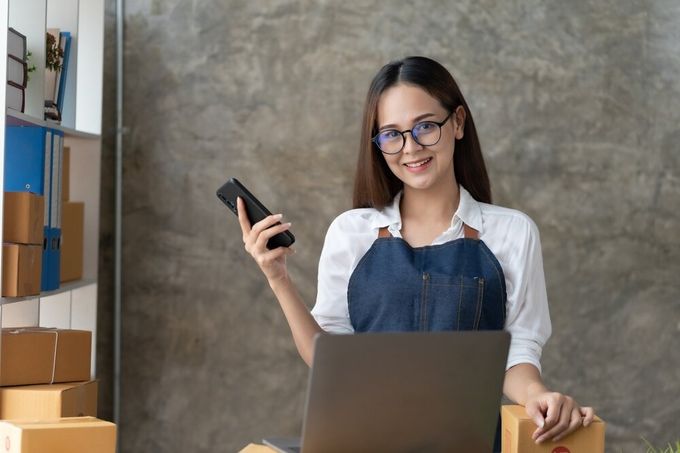 A professional smiling, standing in her office filled with boxes, holding her cellphone in one hand.