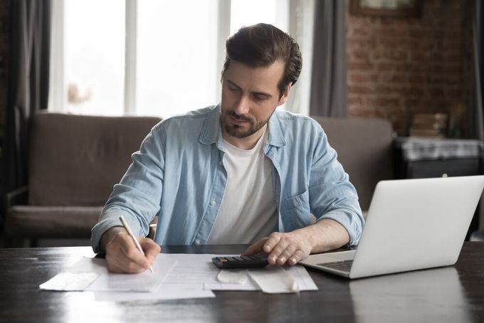 A man seated in his home office, writing something down on paper, with his laptop open next to him.