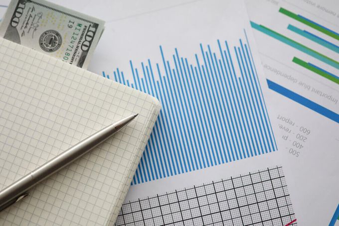 A notebook and a pen placed on a stack of papers, featuring a variety of charts, along with two crisp $100 notes.