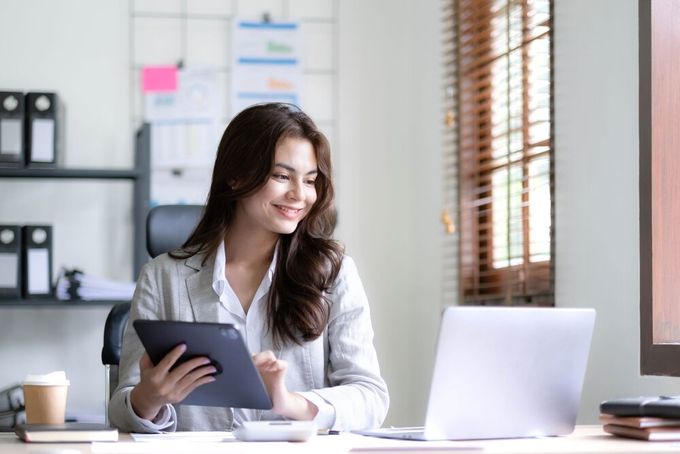 An online store owner smiling, seated in her office, working on her tablet, while looking at something on her laptop.