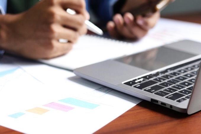 A person seated in front of their laptop, with a pen in their one hand and a phone in the other, with sheets of paper featuring charts on the table below them.
