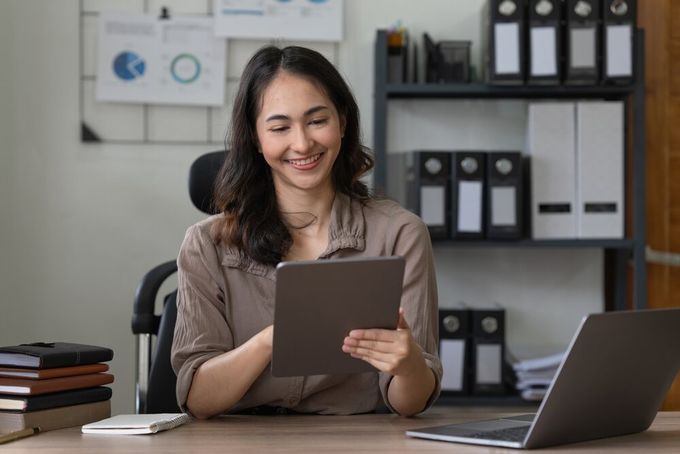 A woman smiling as she works on her tablet in her office, which features a laptop, stack of notebooks, whiteboard on the wall, as well as a book case containing various ring binder files.