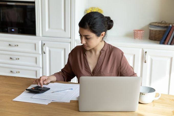 A woman seated in her kitchen, doing calculations with her laptop, various papers, and a coffee mug visible on the table.