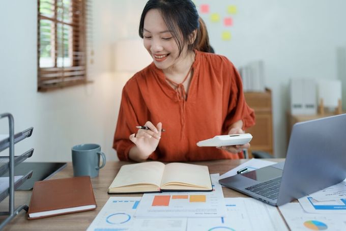 A woman seated at her desk in her home office, holding a calculator in one hand, with two notebooks, her laptop, various pages featuring charts, as well as a cup of coffee visible on the table.