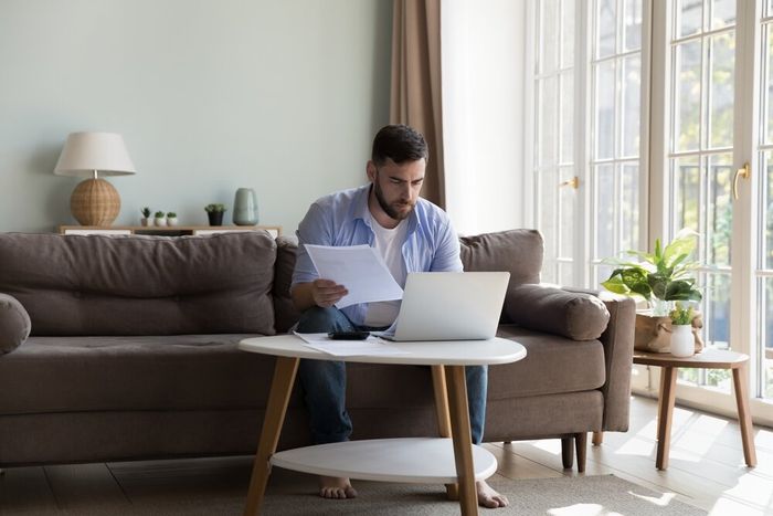 A young man seated on the couch in his living room, holding a stack of papers while he works on his laptop. 