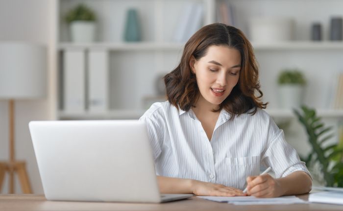 Woman writing on paper in front of laptop at desk