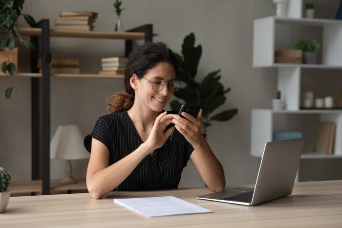 A woman seated at her living room table, smiling at something on her phone, with her laptop and a stack of papers placed in front of her.