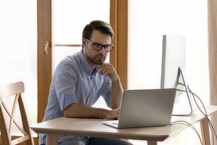 A man seated at a desk in his home office, working on his laptop and a secondary screen.