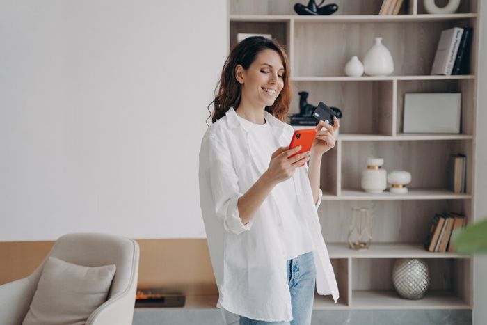 A woman standing in her living room, smiling as she looks at her phone, with a card in the other hand.