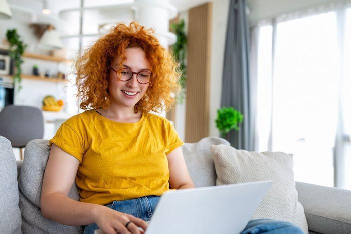 A young woman with red hair, wearing a yellow shirt, seated in her living room, working on her laptop.