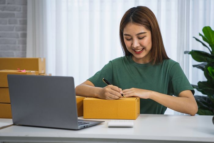 A young woman writing on a packaged product she is shipping to a customer while smiling