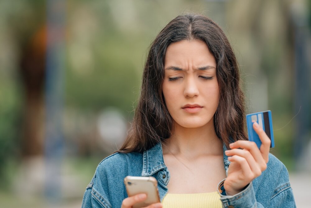 A young woman frowning at the screen of her cellphone while holding her credit card.