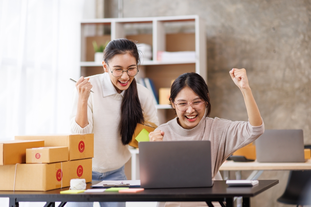 Two women cheering while looking at laptop surrounded by boxes