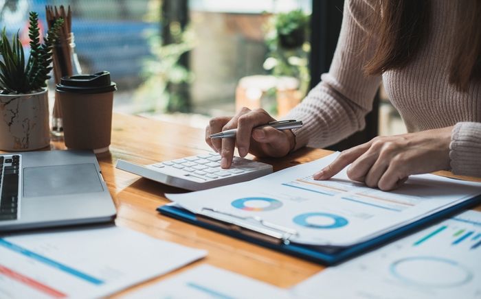 Woman sitting at desk with calculator, laptop, and pages of different graphs preview image