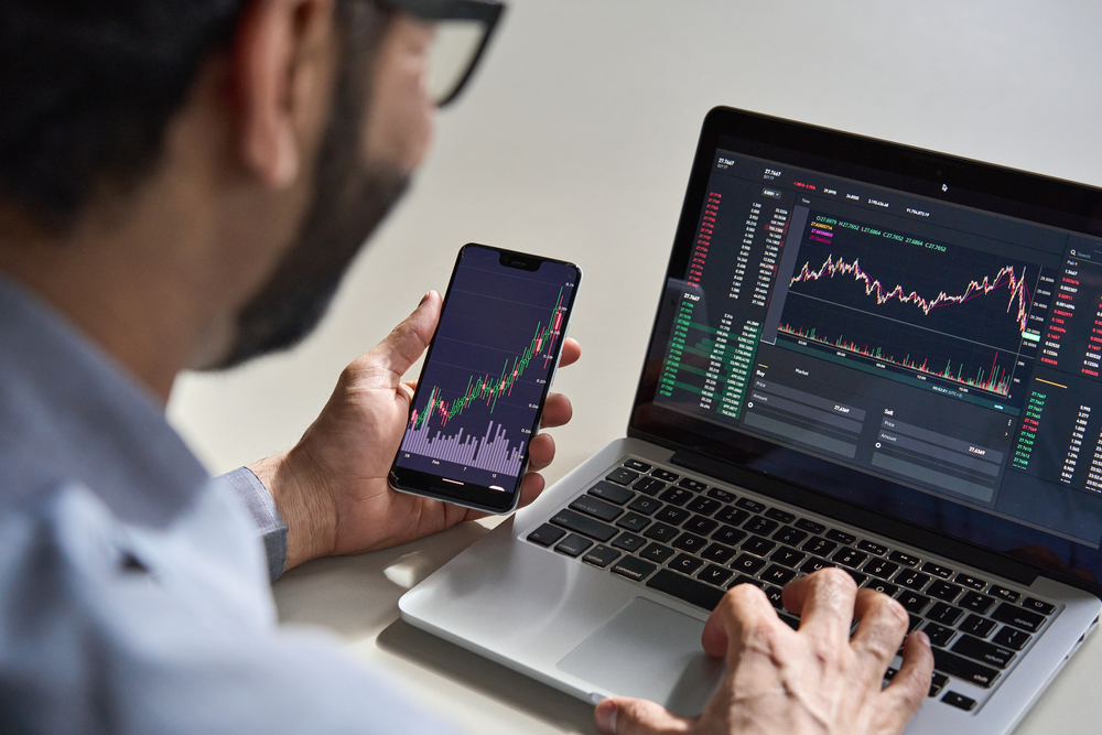 Man looking at stock trading graphs on phone and laptop