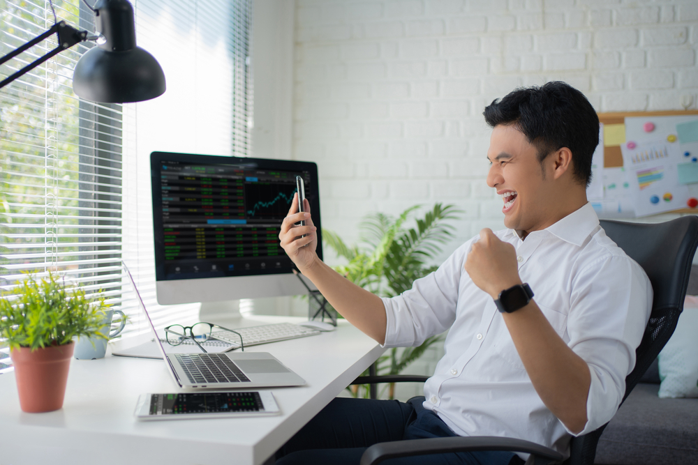 Man smiling at phone with trading stock graphs on PC screen behind him