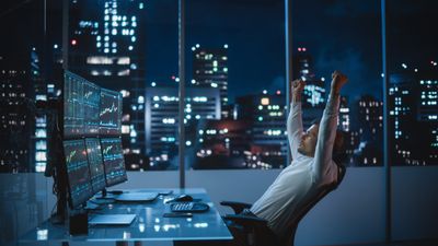 A day trader cheers in front of multiple screens showing trading information on stocks with city buildings in the background