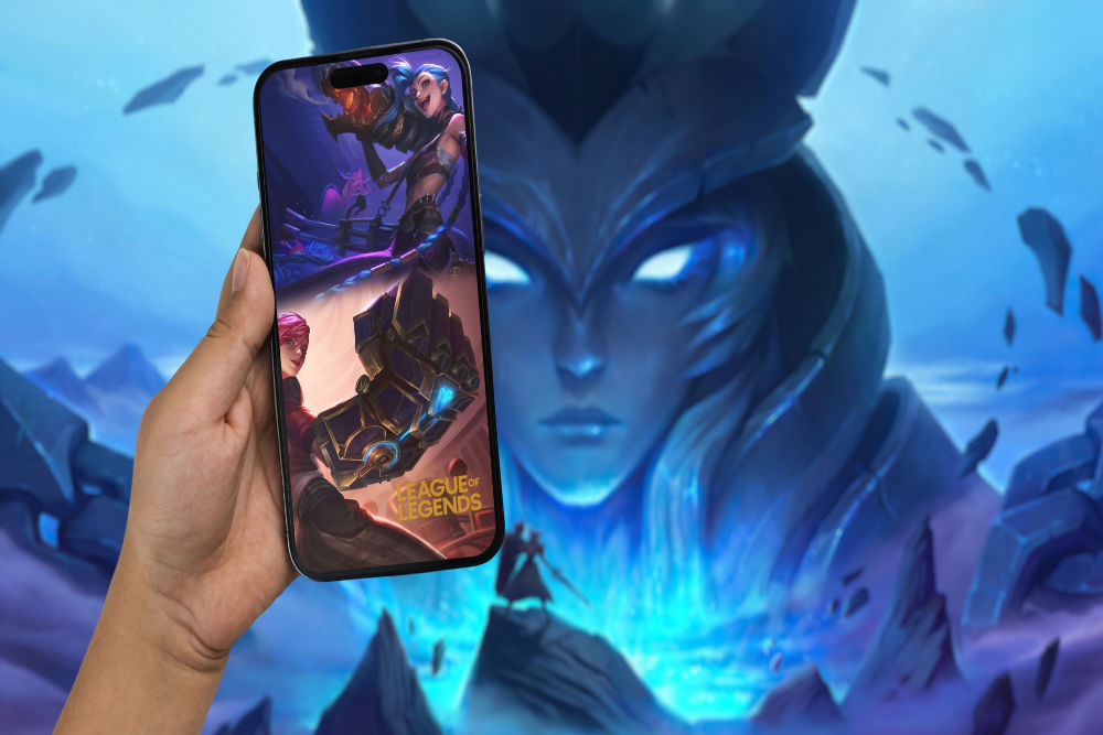 A hand holding a smart phone, with the League of Legends logo and the character 'Jinx' displayed on the screen, against a background featuring a still from the game.