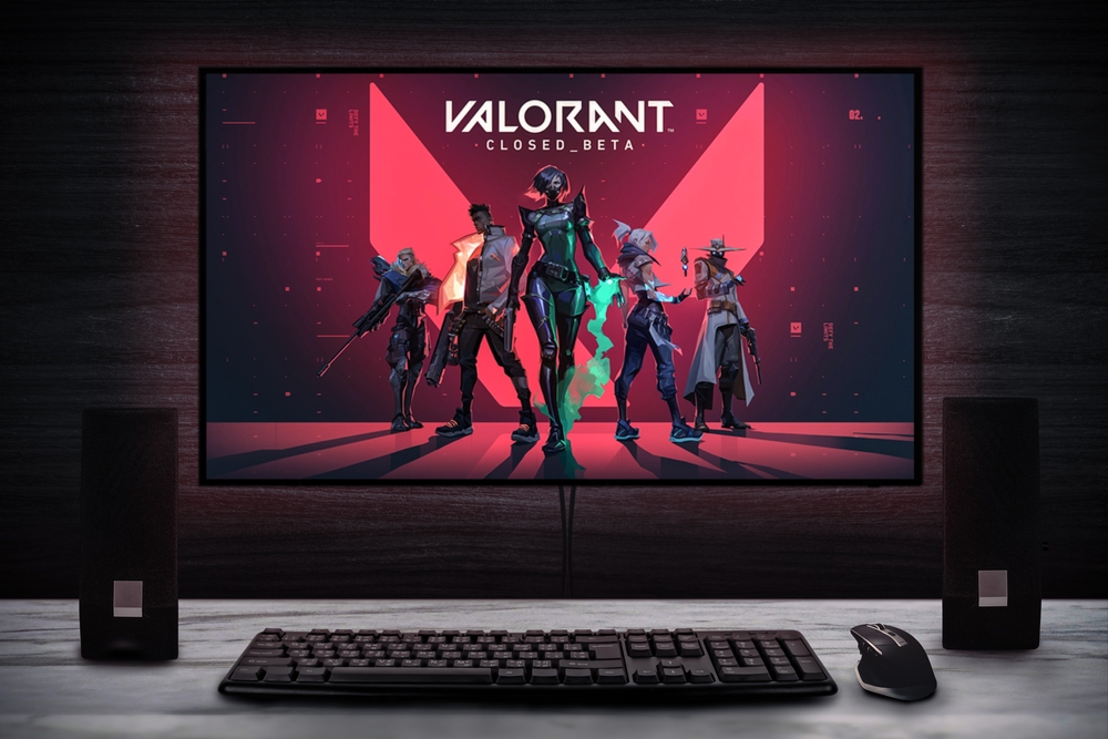 Gaming PC setup displaying Valorant loading screen behind speakers, keyboard, and mouse
