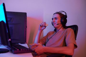 A young man, seated at his desk with a headset on his head, watching something on his computer as he enjoys a snack.