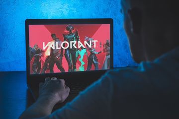 A PC gamer seated with his laptop open, with the screen displaying Valorant characters against a red background, with the game's name displayed in the forefront.