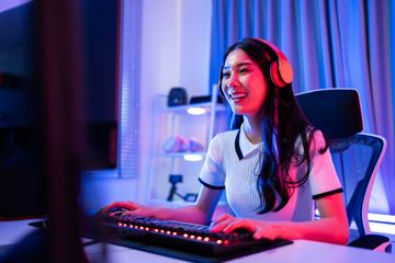 A young girl seated at her desk in a room lit with blue and purple LEDs, working on her computer with a headset on her head.